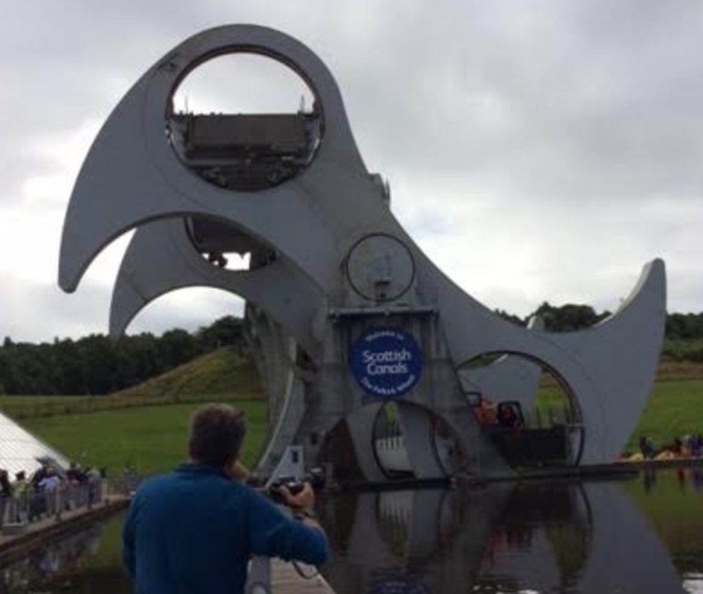 a man taking a picture of a large metal structure with Falkirk Wheel in the background