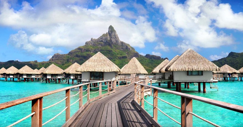 a wooden walkway leading to a small bungalow on water with Bora Bora in the background