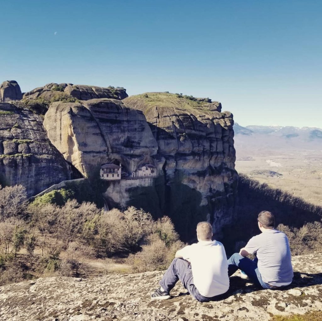 two men sitting on a rock looking at a building on a cliff