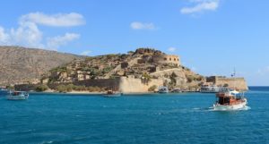 a large stone building on a hill next to a body of water with Spinalonga in the background