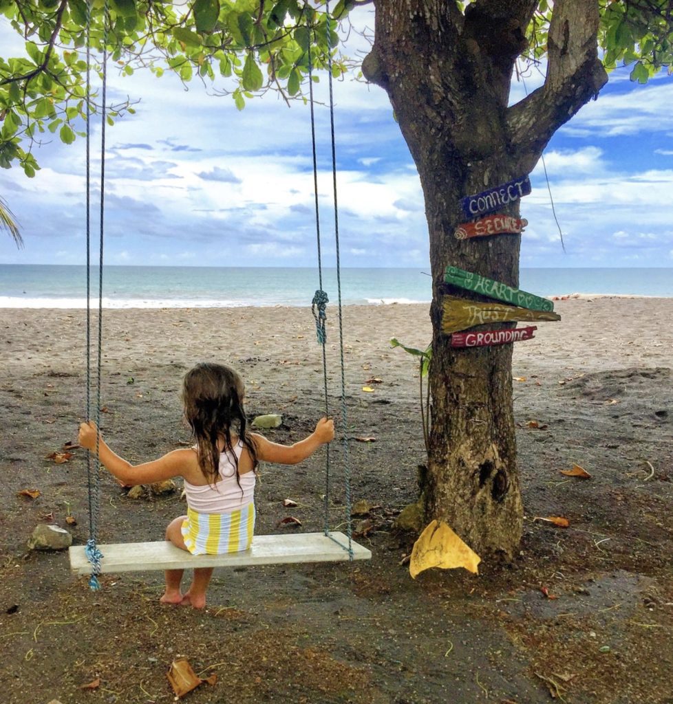 a girl sitting on a swing under a tree on a beach